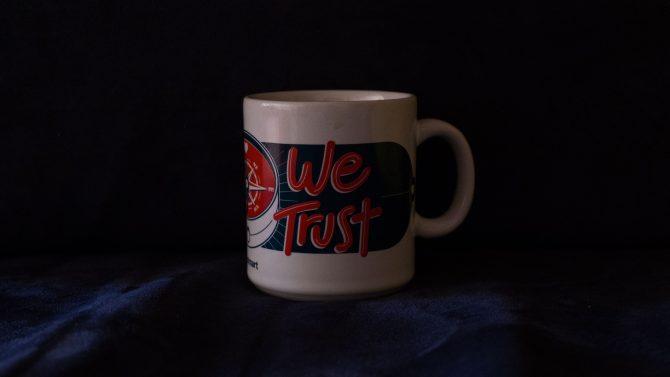 Foto de una taza blanca encima de telas azules cuya frase dice "We trust" en letras rojas.
