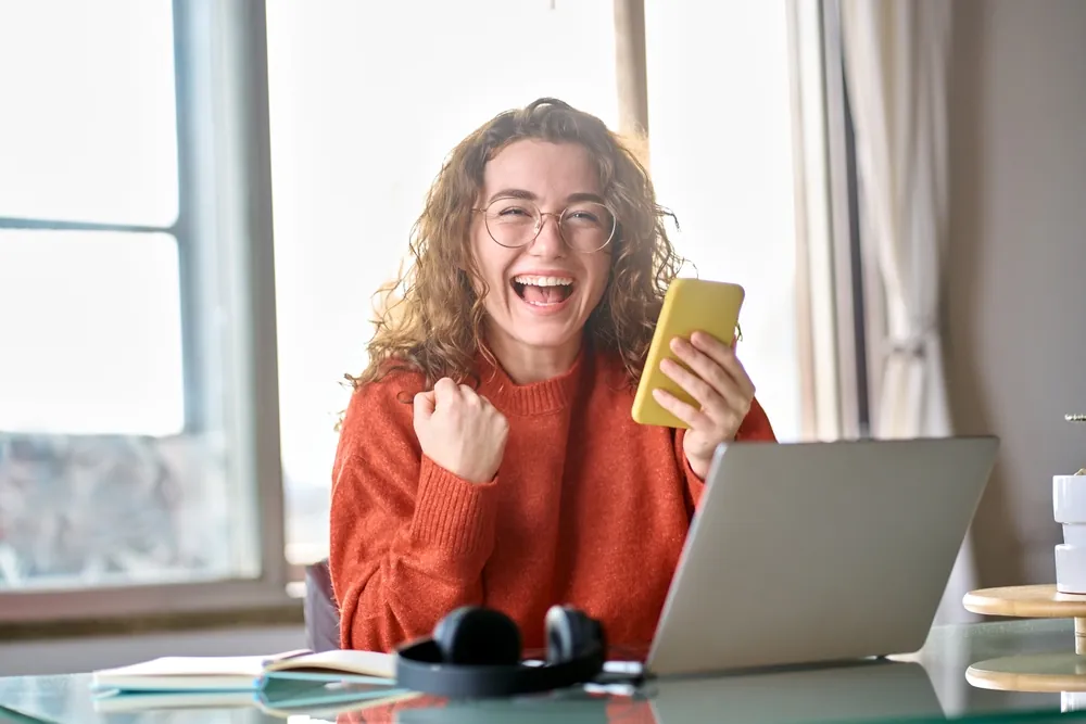 Mujer sonriendo y celebrando con el celular en la mano frente a su laptop, después de vender un infoproducto