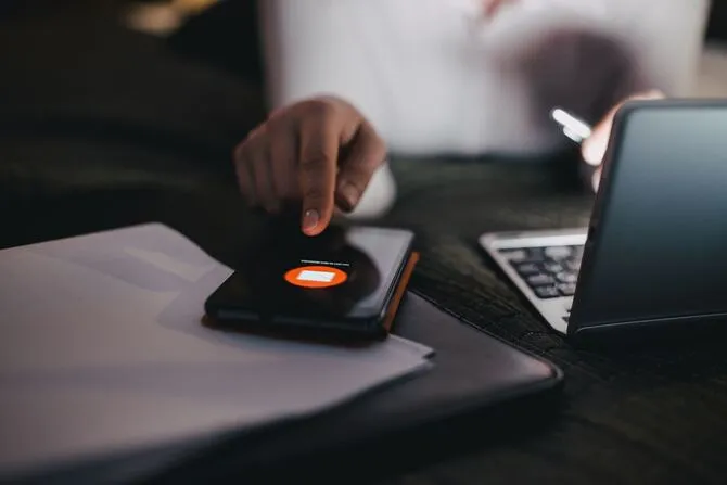 Person tapping a smartphone showing a new email notification, next to a laptop and papers on a desk