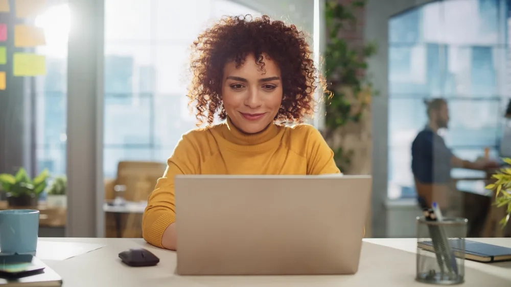 Smiling woman working on a laptop in a bright office, using Hotmart tools to sell digital products