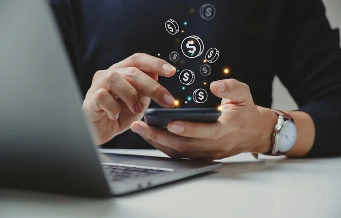 Persona usando un celular junto a una laptop con íconos de monedas flotando, que representan la monetización en Instagram