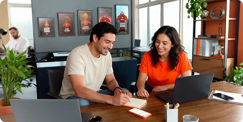 Dos compañeros de trabajo revisan una libreta y sus laptops en una oficina cálida, escena que ilustra el acompañamiento a los negocios digitales.