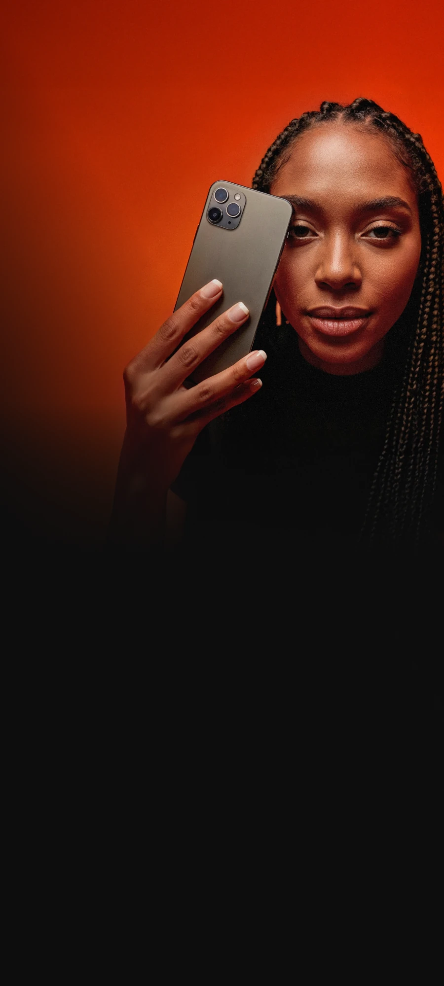 No centro da imagem há uma jovem mulher com pele escura e cabelo com tranças, usando uma blusa preta, segurando um smartphone cinza na frente do rosto, com expressão neutra e fundo laranja e marrom em degradê